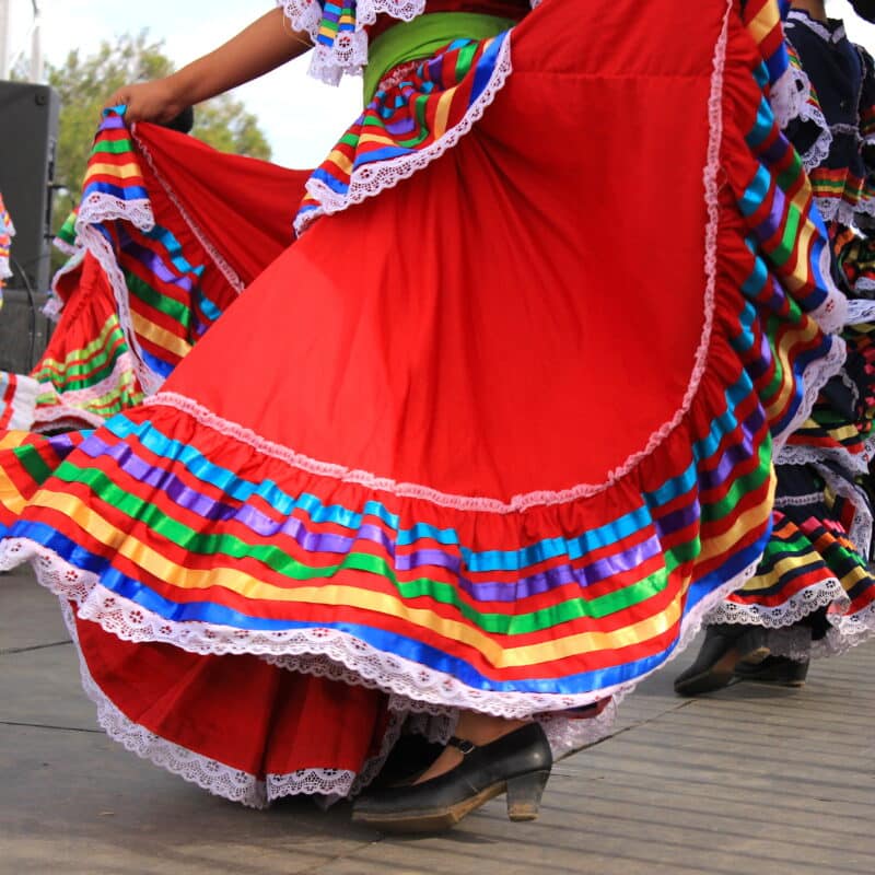 Dancers in bright traditional folklorico dresses with rainbow striped ruffles swirling during a performance in an outdoor setting.
