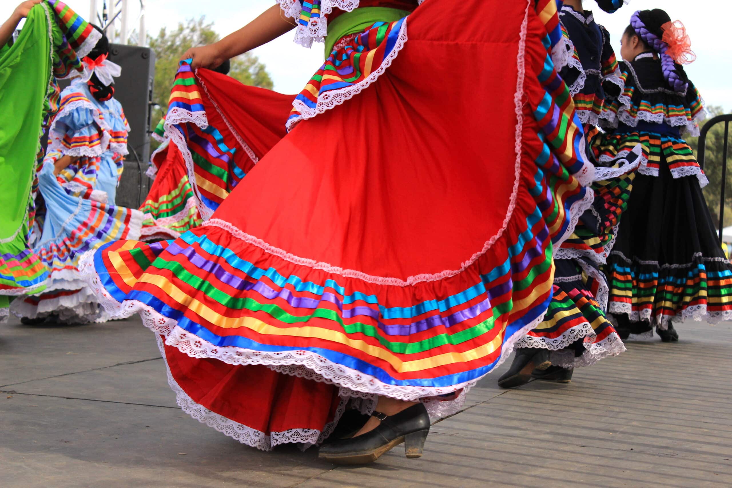 Dancers in bright traditional folklorico dresses with rainbow striped ruffles swirling during a performance in an outdoor setting.