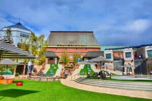 Outdoor shopping plaza with green artificial turf, striped umbrellas, and a building sign reading The Gateway under a blue sky.