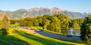 Park scene with a row of parked cars, green lawn, trees, and a lake with a fountain, mountains in the distance under a clear blue sky