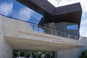Exterior view of a modern concrete and glass museum entrance with copper-tone overhangs and large windows reflecting the sky; sign reads 'Natural History Museum of Utah' (partial).