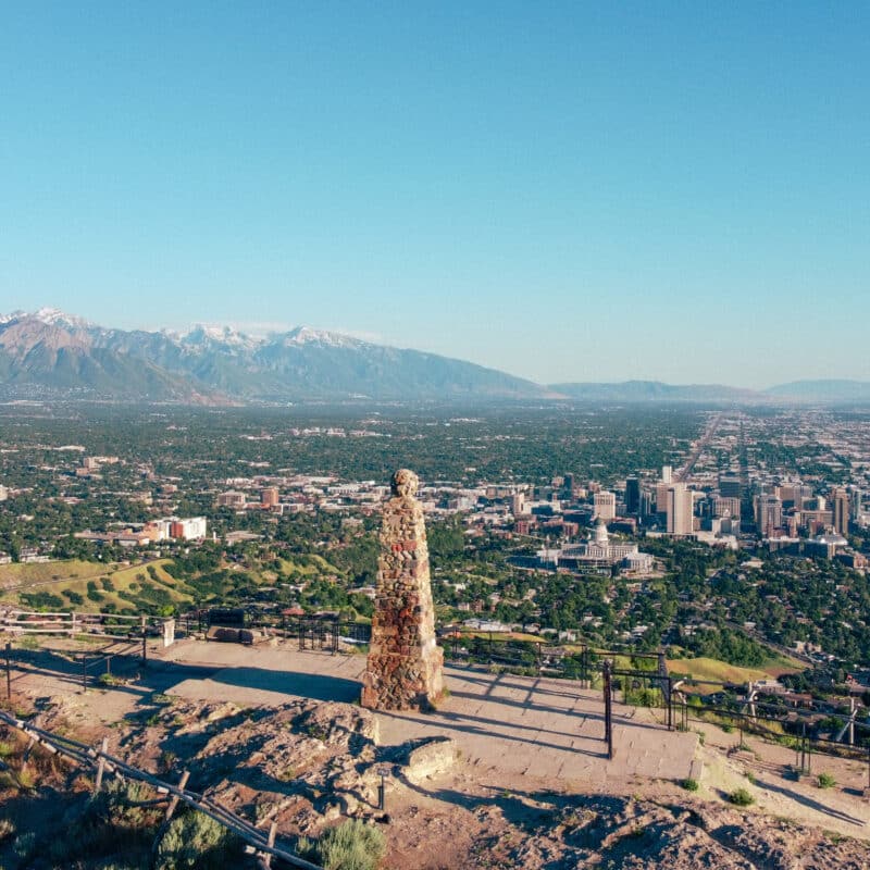 Panoramic view of a city and distant mountains from a hilltop, with a stone pillar monument in the foreground.