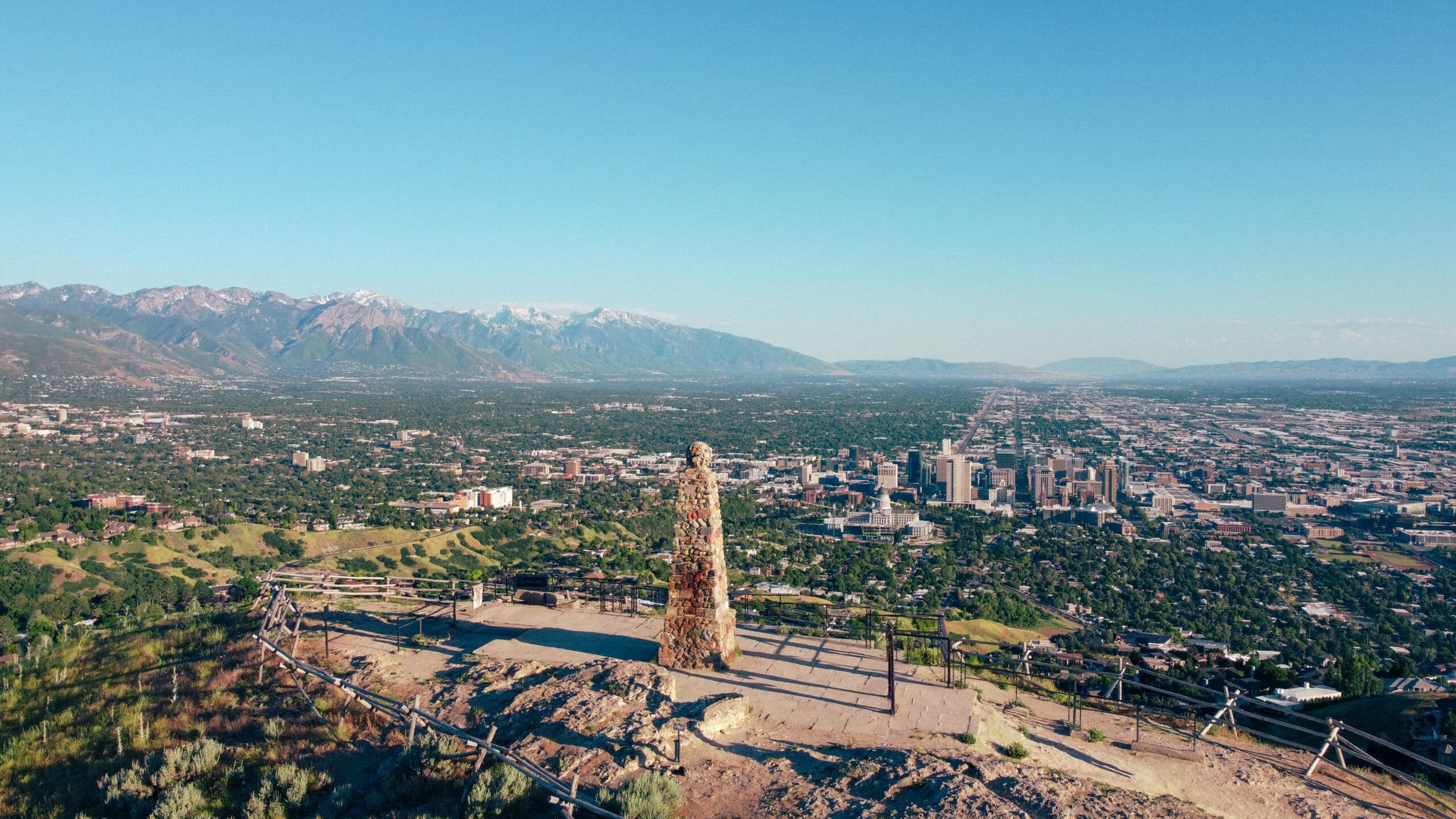 Panoramic view of a city and distant mountains from a hilltop, with a stone pillar monument in the foreground.