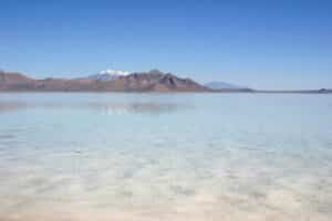 Wide landscape of a shallow, pale turquoise salt lake with distant brown mountains and snow-capped peaks under a clear blue sky; calm reflection on water surface.