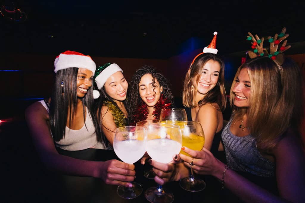 Group of five young women in festive hats clinking cocktails at a holiday party in a dim club setting
