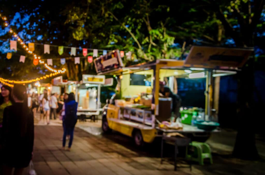 Blurred night market scene with string lights, food stalls, and people walking along a paved path.