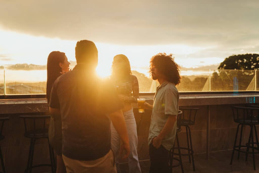 Friends chatting on a rooftop terrace at sunset, holding drinks with a glass railing and warm backlighting behind them.