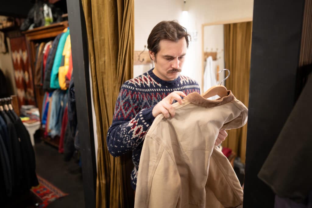 Man wearing a blue patterned sweater examines beige pants on a hanger in a clothing store. The shop has jackets on racks around him.
