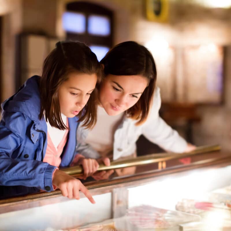 Two girls lean over a glass display case, pointing and closely examining items inside a softly lit museum or shop.