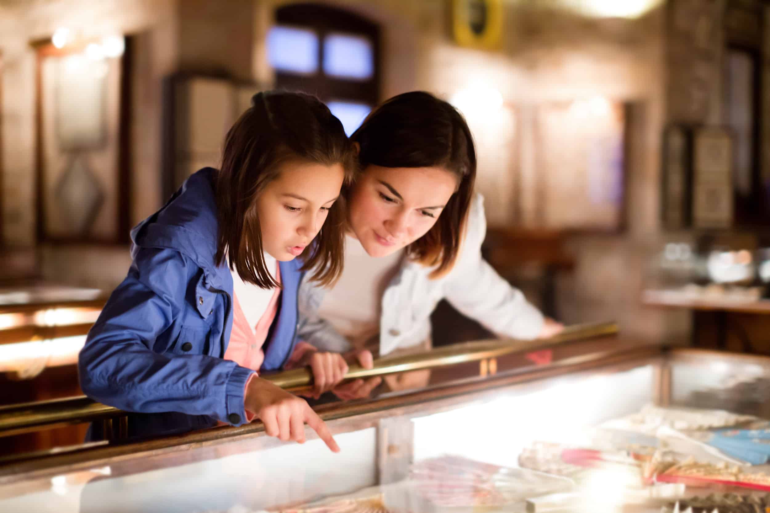 Two girls lean over a glass display case, pointing and closely examining items inside a softly lit museum or shop.