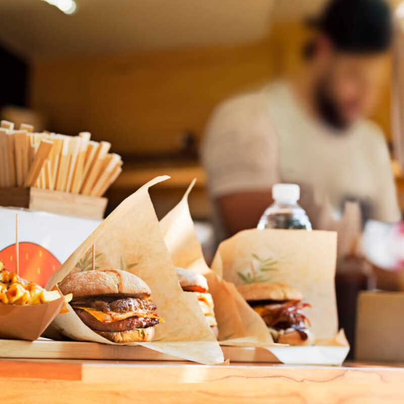 Close-up of burgers and fries on a wooden counter, with a green-topped hot sauce bottle in the background at a fast-casual restaurant.