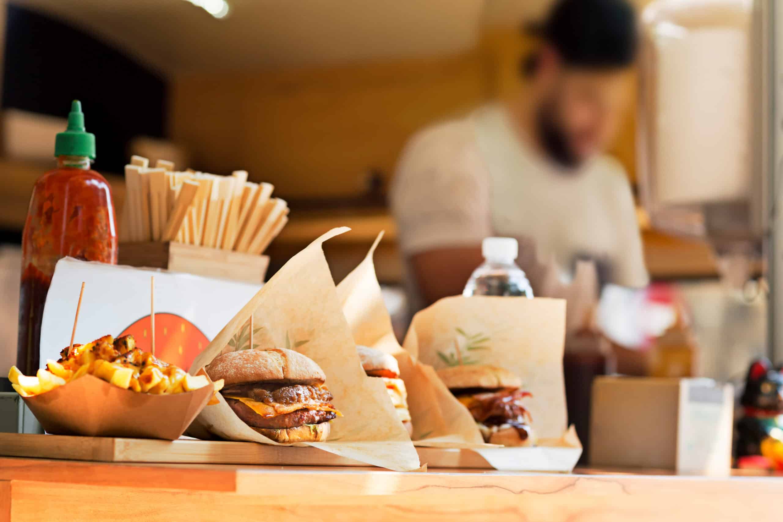 Close-up of burgers and fries on a wooden counter, with a green-topped hot sauce bottle in the background at a fast-casual restaurant.