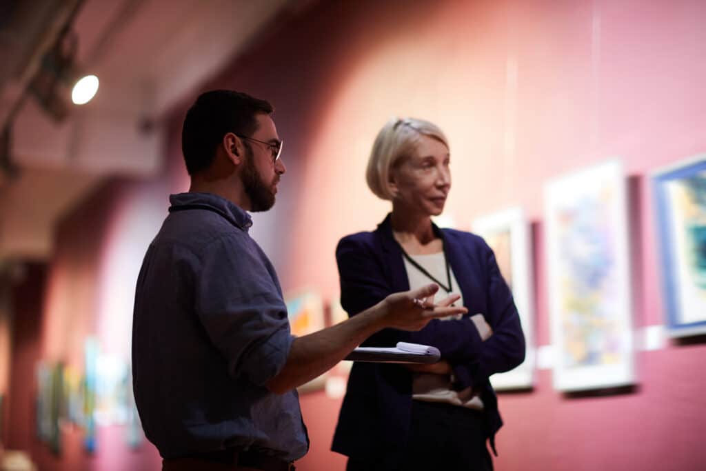 Two professionals talking in a gallery or museum hall; man with beard and glasses facing left, woman in blazer listening with a clipboard nearby.