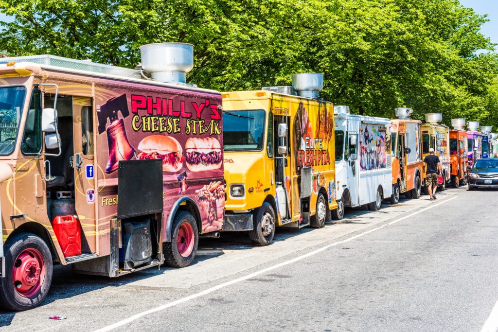 Row of colorful food trucks parked along a street with green trees overhead, signage visible on the sides (Philly's Cheese Steak, etc.).