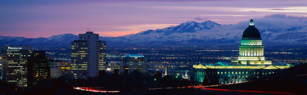 Evening city skyline with a domed government building illuminated in green and snow-capped mountains in the distance at dusk.