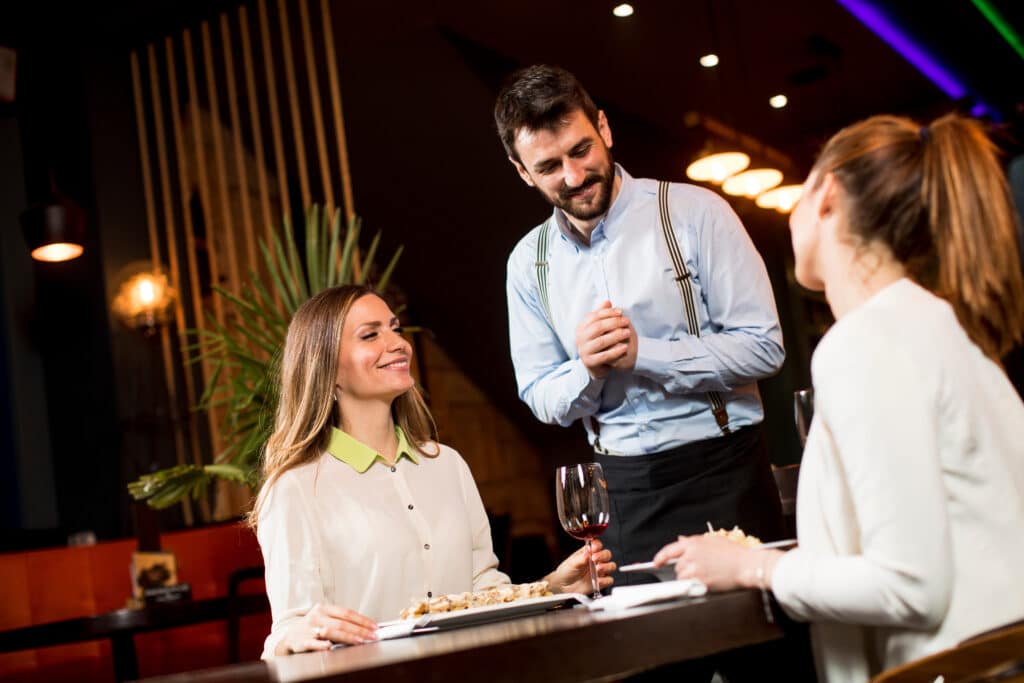 Woman wearing a light blouse with a lime collar smiles as a waiter in suspenders chats with two diners at a dimly lit restaurant bar, a wine glass and appetizers on the counter nearby.
