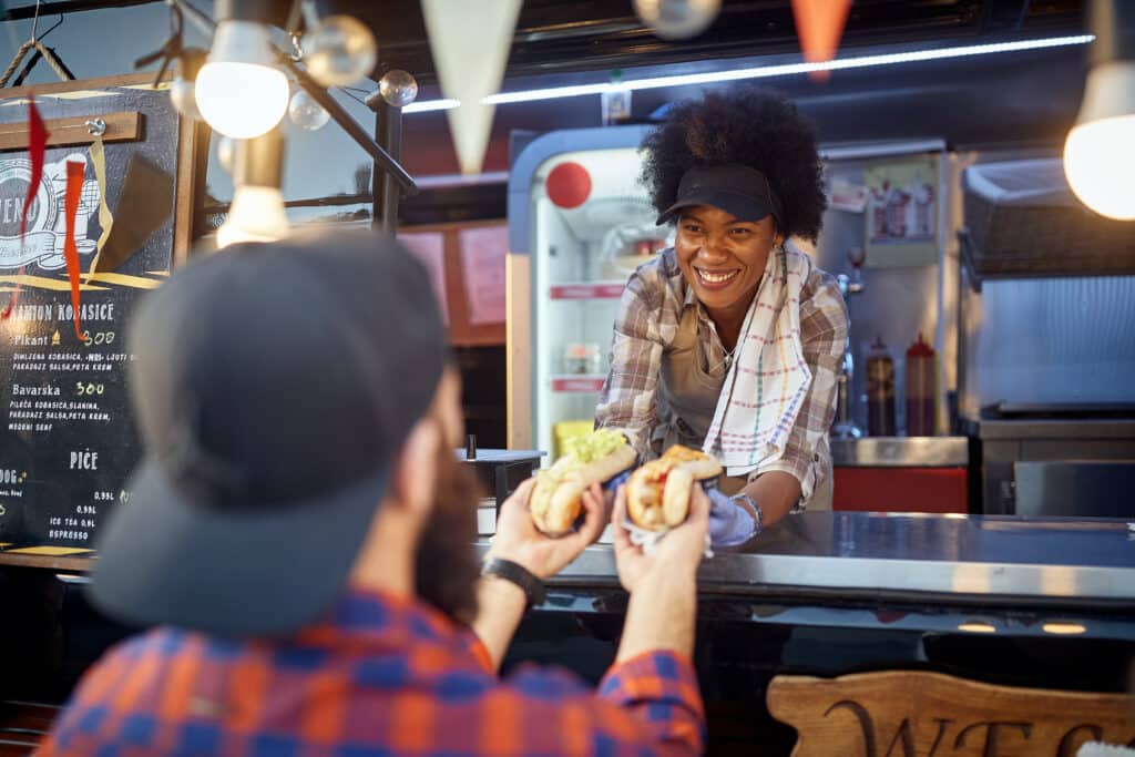 Smiling female street vendor hands hot dogs to customers at a counter with a chalkboard menu nearby.