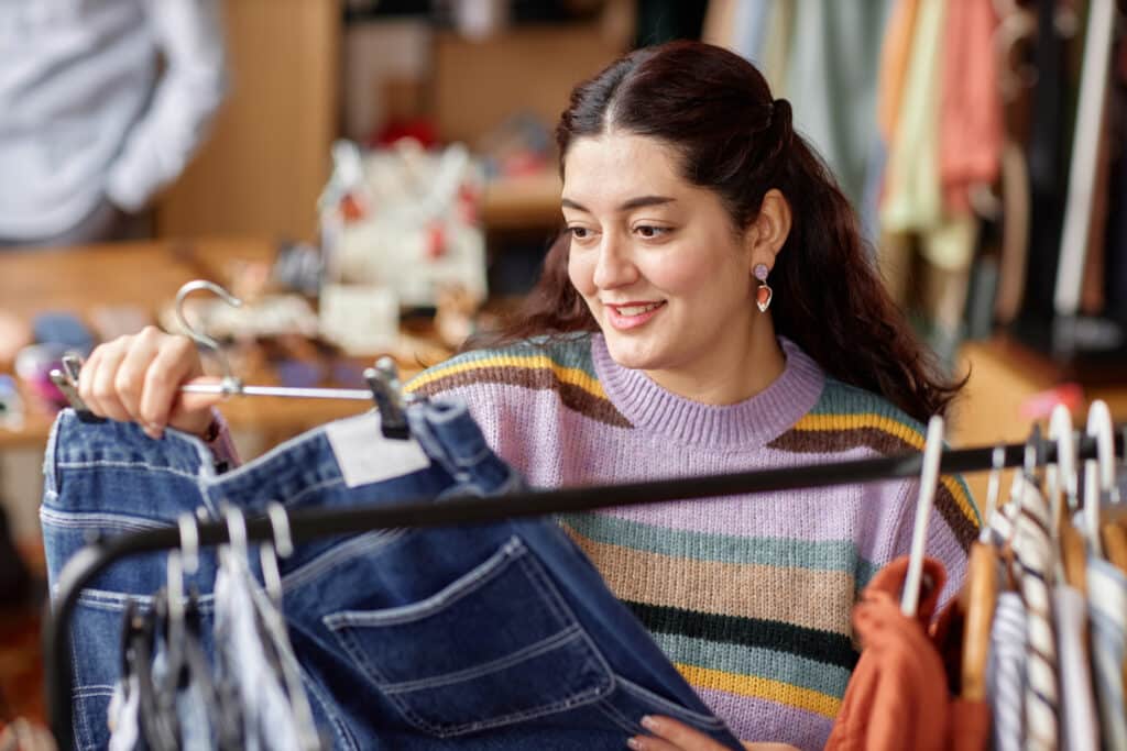 Woman in a striped sweater browses denim jeans on a clothing rack in a shop, smiling as she looks at the jeans.