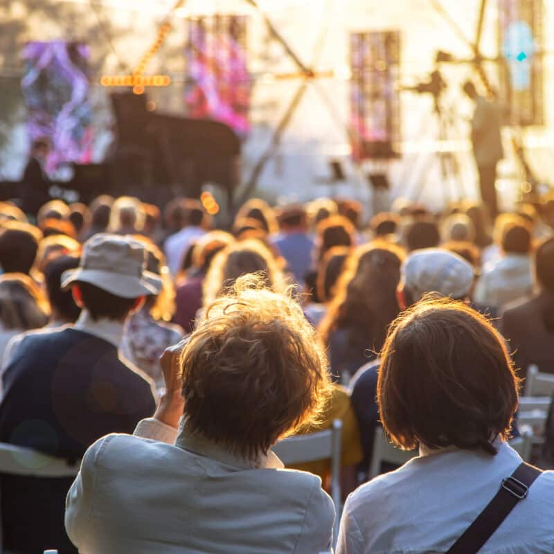 Crowd seated outdoors at a sunny concert or festival with a stage in the distance and warm light.