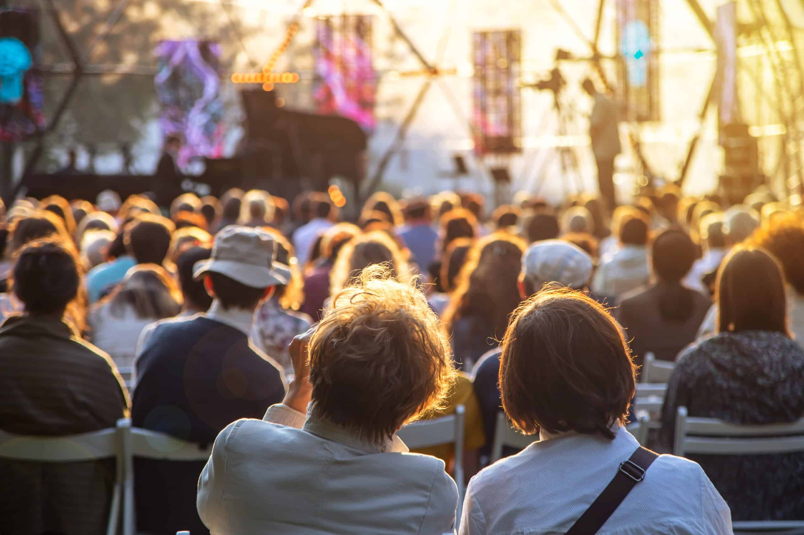 Crowd seated outdoors at a sunny concert or festival with a stage in the distance and warm light.