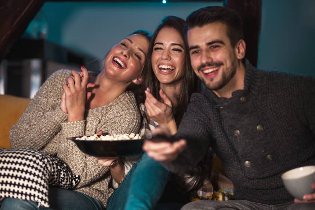 Three friends laughing together on a couch, sharing a bowl of popcorn in cozy sweaters at a casual gathering