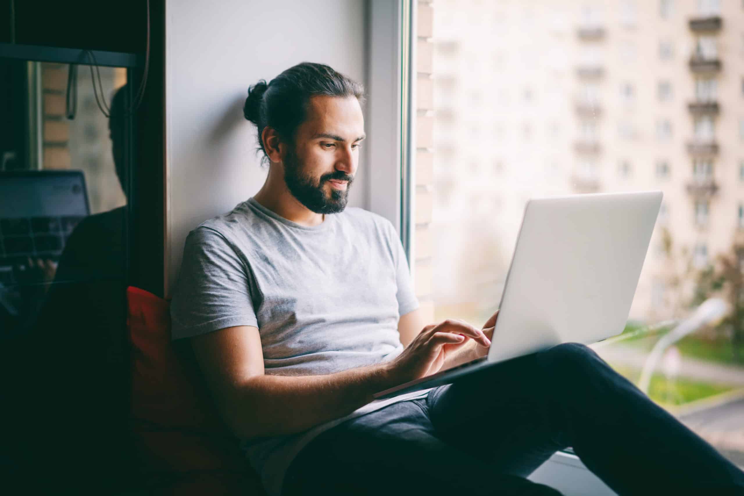 Man with a beard and bun sits by a window, typing on a laptop.
