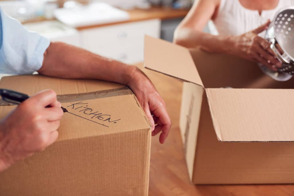 People packing cardboard boxes, one person writing with a marker while another caps a box with a metal lid nearby (moving/packing scene).