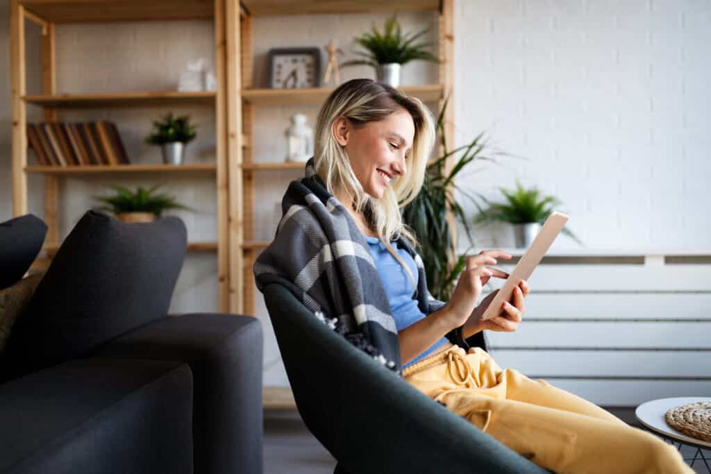 Woman smiling while using a tablet on a sofa in a cozy living room with wooden shelves and houseplants behind her